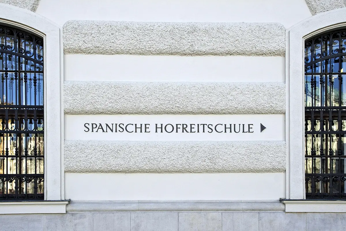 A dark metal sign reading Spanische Hofreitschule with an arrow, on a pale rendered wall between two barred arched windows