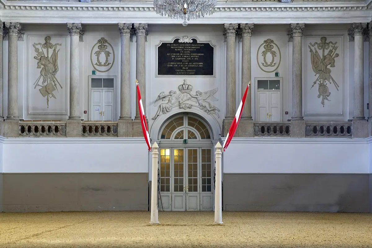 The riding entrance of the Winter Riding School, with a Latin inscription plaque, two angels holding a crown above a glazed arched door, two white posts with Austrian flags and a sand floor